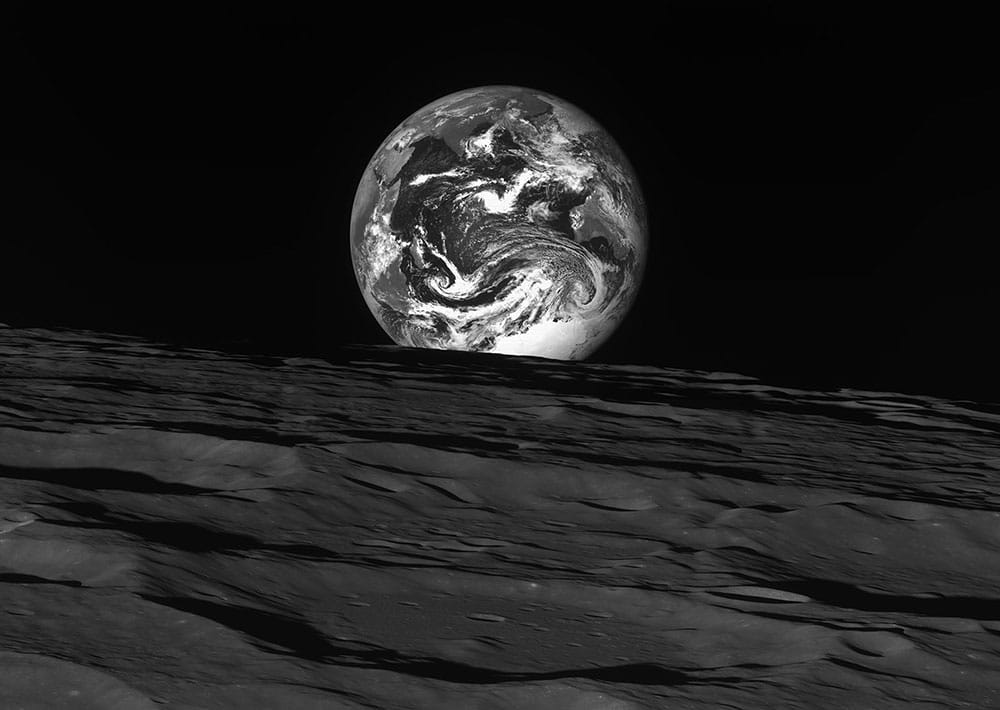 Our Earth is seen rising above the Moon’s horizon in this sharp black & white image captured by South Korea’s KPLO lunar orbiter. The lunar surface in front shows large craters spanning the scene.